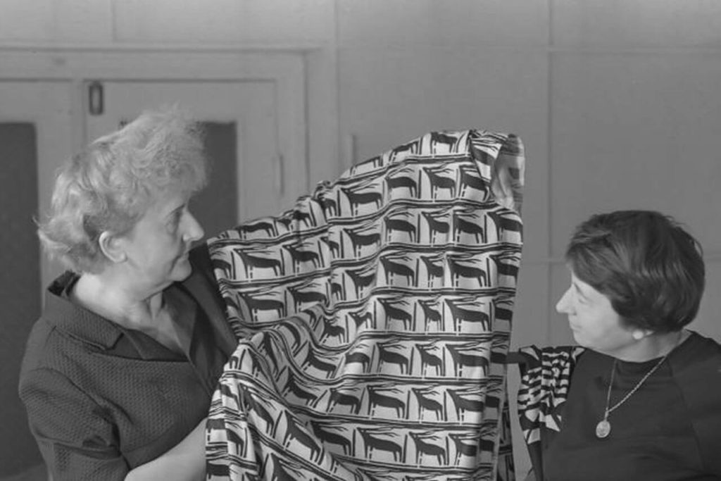 Two elderly women are shown in a black and white photograph: the one on the left, with grey hair, is presenting a white cloth with a black design to the woman on the right, who has black hair.