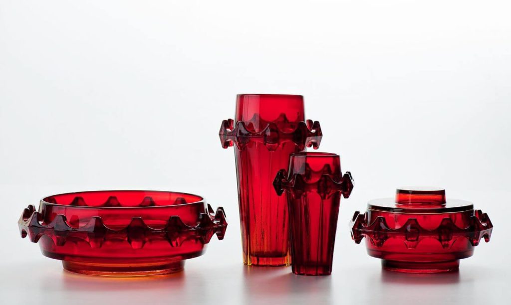 A set of red glassware. Four circular-plan vessels: a sugar bowl, two vases, and an ashtray. All have a circular, rhomboid shape with a sculpted ornamented line around each.
