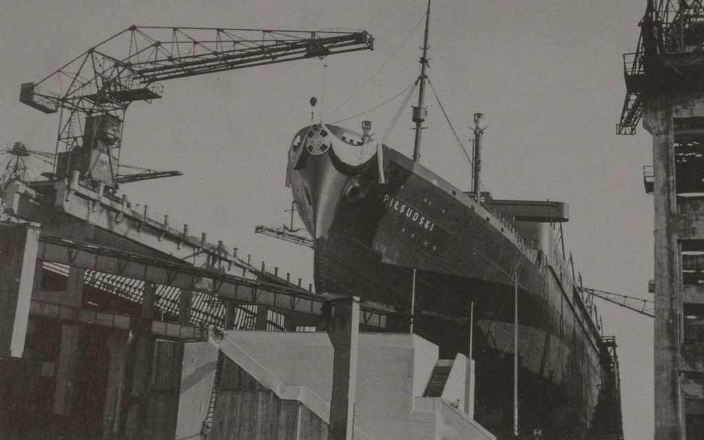 A monochromatic photograph of an enormous ship in the dockyard. Its corpus is black with the white inscription 'PIŁSUDSKI' visible on the right side. The prow is adorned with a white eagle and a cross symbol. Above the ship, some cranes can be seen.