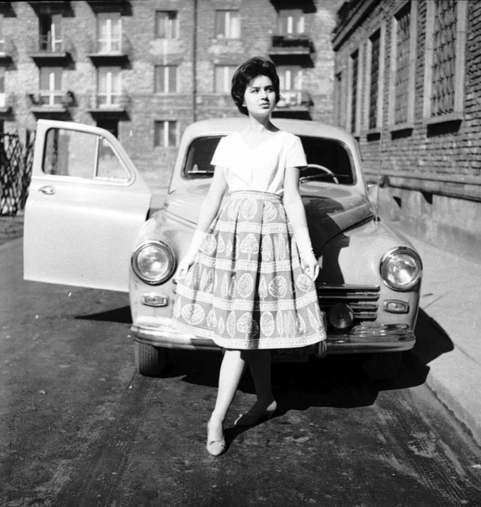 "A black-and-white snapshot depicts a young black-haired woman dressed in a white blouse, white long gloves, and a tree-patterned medium-sized skirt. She's leaning against the front bumper of a Warszawa car. "