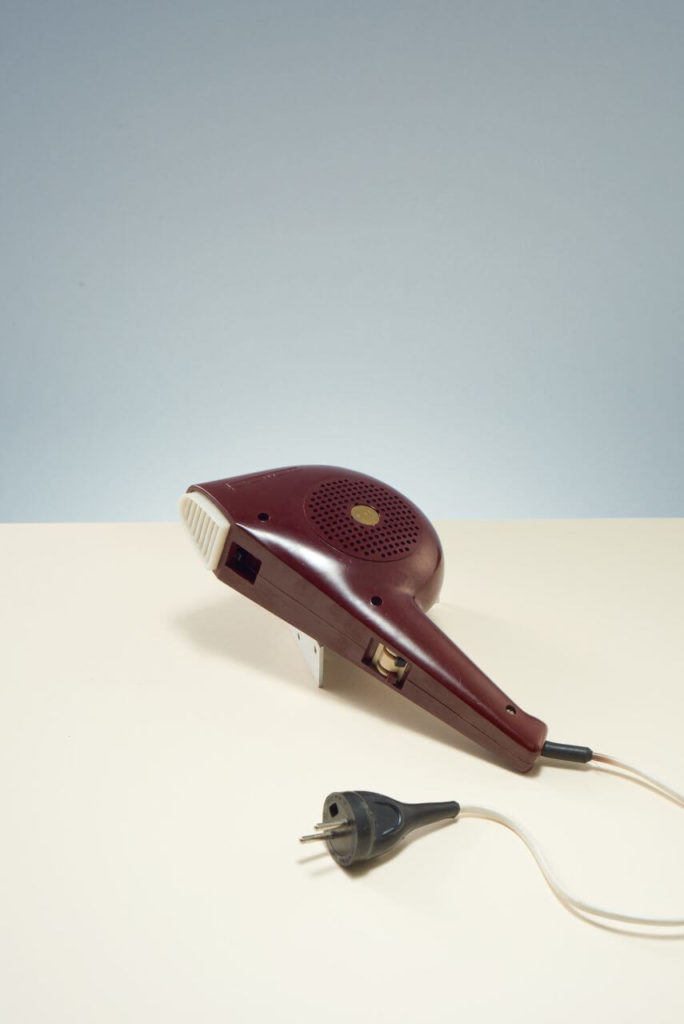 A small hand-held hair dryer. It is mostly brown. It has a white cord and a black plug. Its main vent is white. The body has small ventilation holes.
