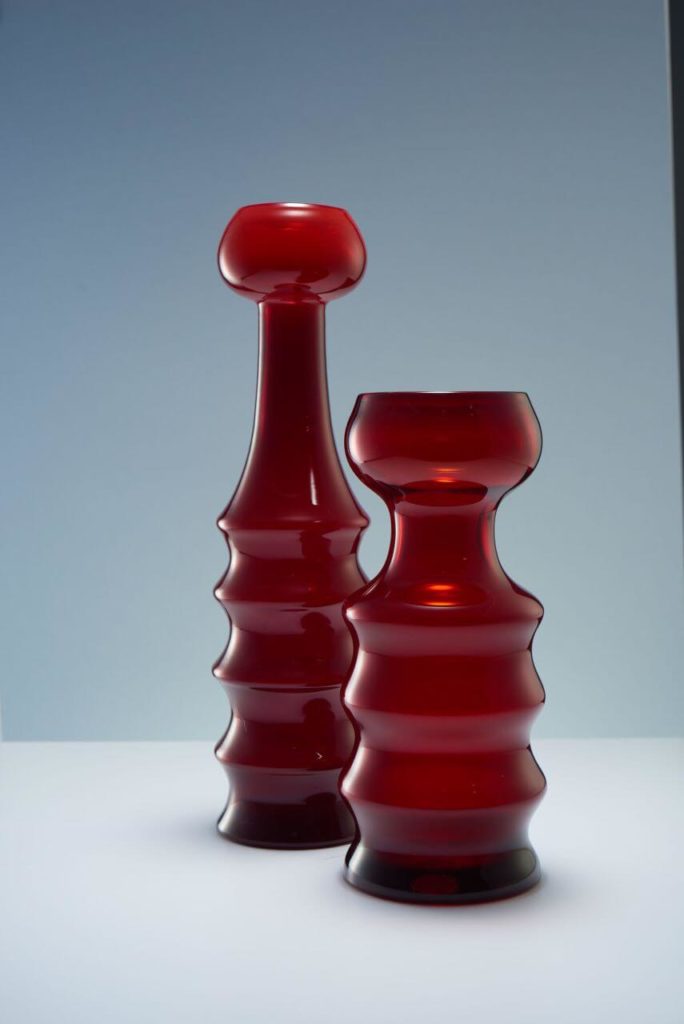 Two red translucent glass vases. The lower parts of both are segmented. The neck of the vessel on the left is thin and elongated like the neck of a giraffe, while the object on the right has a short and massive neck.