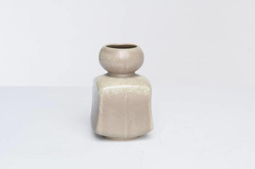A small ceramic vase, shaped like an inkwell. Its lower segment is square, and the upper segment is a rounded bowl. Its colour is light, heterogeneous and resembles grated sandstone.