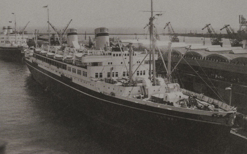 A snapshot of a massive transatlantic ship near the shipyard in black and white. The corpus is black with white characters spelling out 'BATORY.' Three chimneys tower above the deck, which is white.