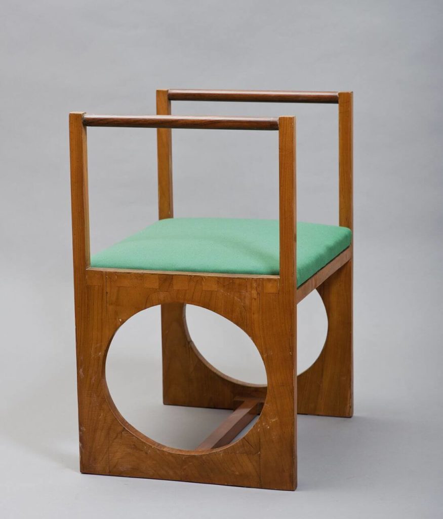 "A bedroom stool that consists of two wooden, light-brown, square-shaped frames. In between, there is a green, plush seat. The base is made of two massive wooden legs with a prominent circular cutout in the middle. Both are joined by a beam at the bottom of the structure. "