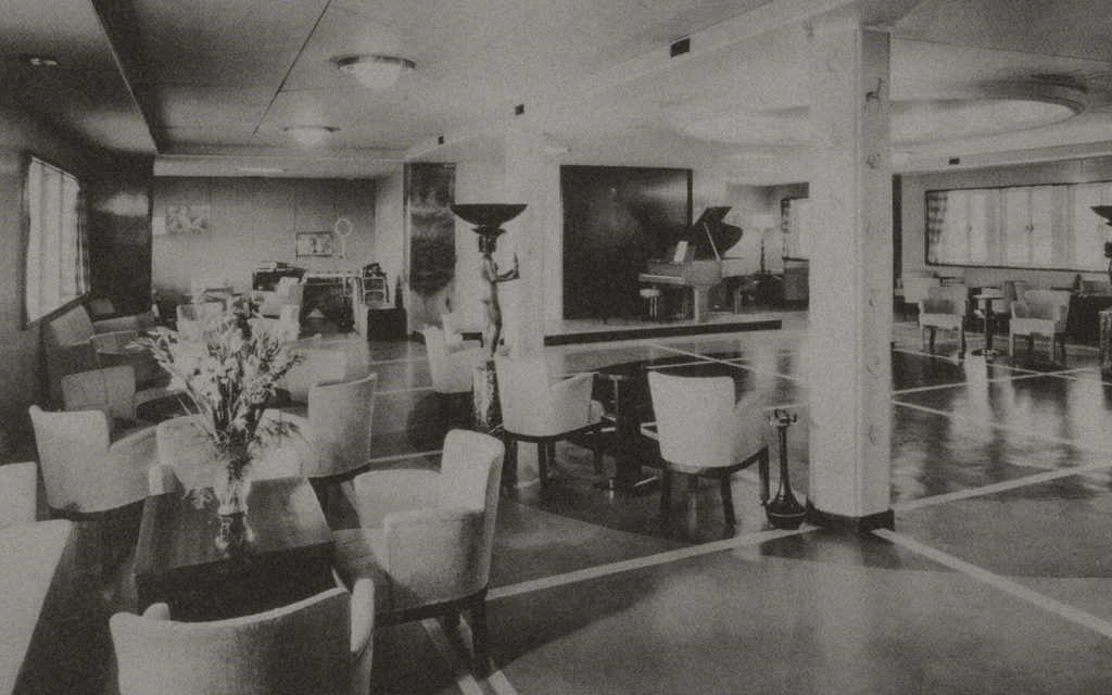A black and white photo of a ship's salon. The room is sparse. On the left, there are some tables with flowers and surrounded by chairs. On the right, the black grand piano is visible, as is an empty space-probably for dancing. In the middle, a statue of a nude woman holding a big vase on her head is placed.