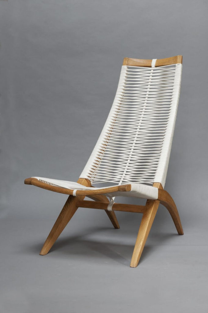 An armchair with wooden legs and a high, slender backrest, which, together with the seat, is made of white threaded tape-like material.