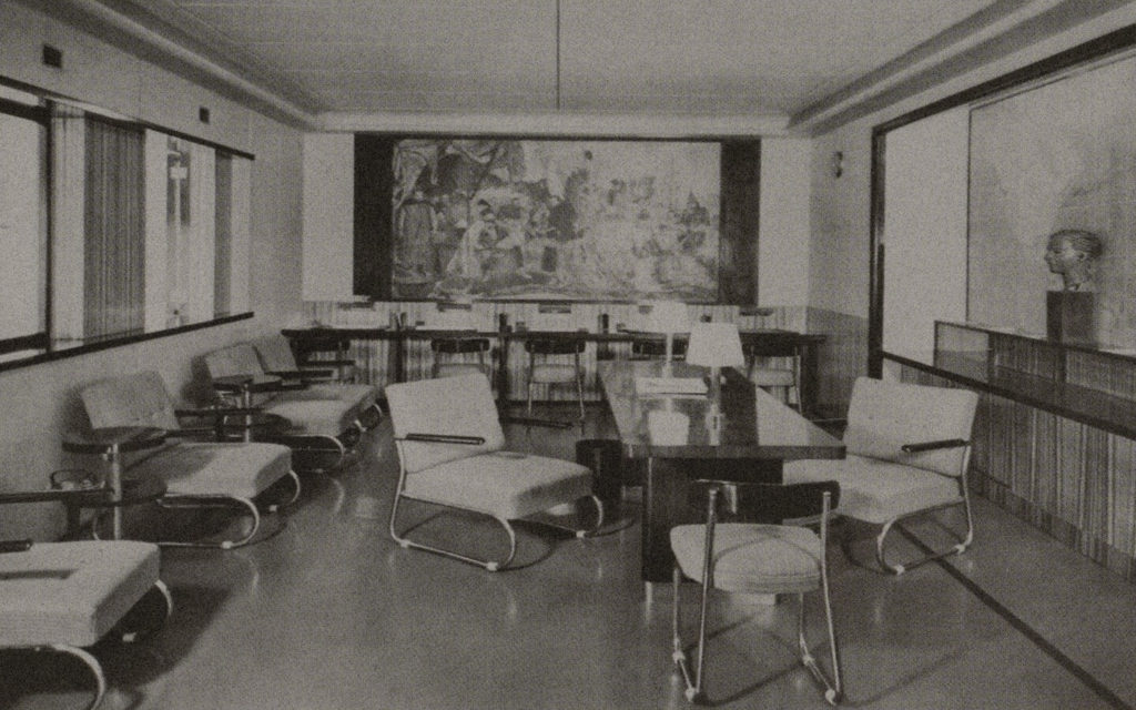 A snapshot of a ship's reading area in black and white. There are numerous tables with lamps adorning them, as well as cozy armchairs. A large replica of a historical painting hangs on the back wall. A statue of a human head can be seen below the right-hand wall.