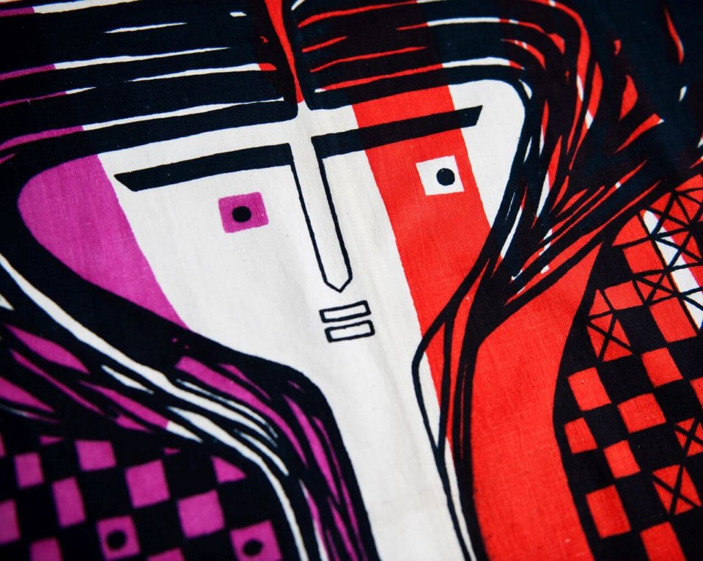 A textile pattern showing a stylised female head on a red, pink and white striped background. The head has a triangular shape with thick eyebrows, straight nose, two black dots for eyes, two small rectangular lips and thick black lines for hair outlining the head.
