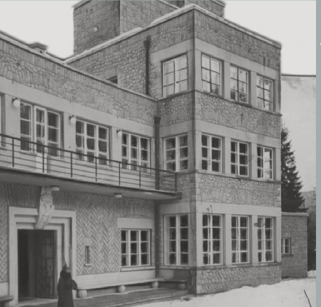 A black and white photograph made in winter. It depicts a section of a villa with flat roofs and simple geometrical patterns. The building's right side has three stories, whereas the left side has only two. Above the entryway, a balustrade may be seen. A woman dressed in a fur coat stands nearby.