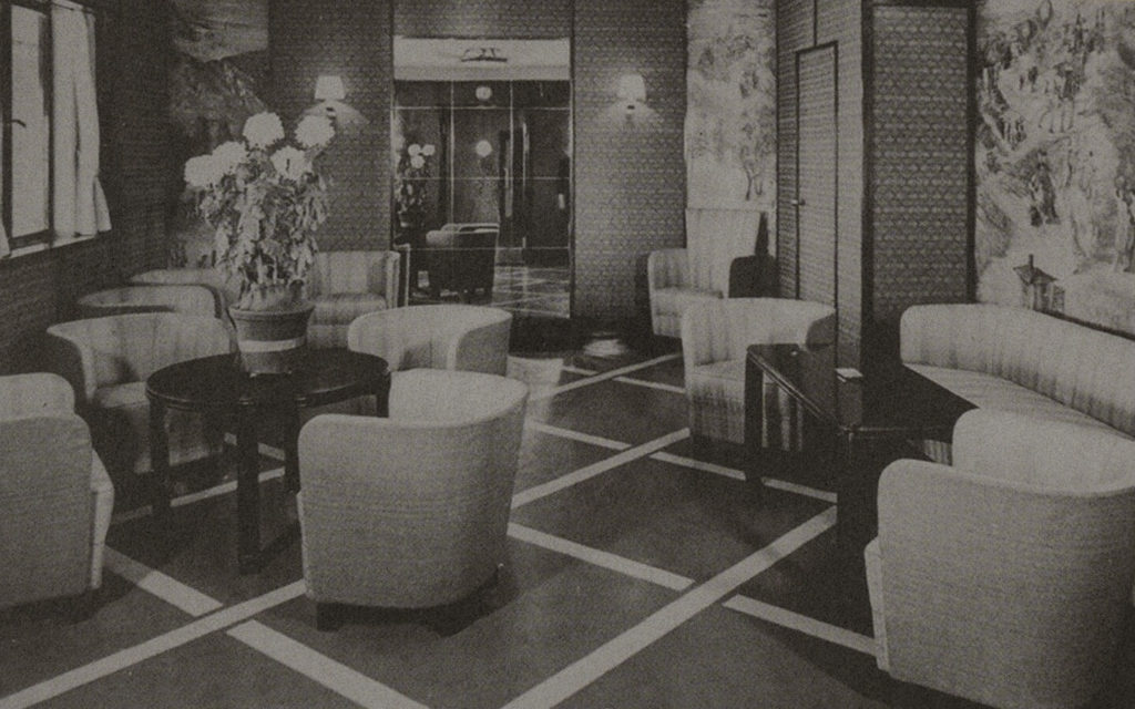 A black-and-white snapshot of a room with comfortable armchairs, a round black table on the right with a conspicuous potted flower, and a rectangular table on the left. The back wall is adorned with mirrors, while the left-hand wall is decorated with a tapestry.
