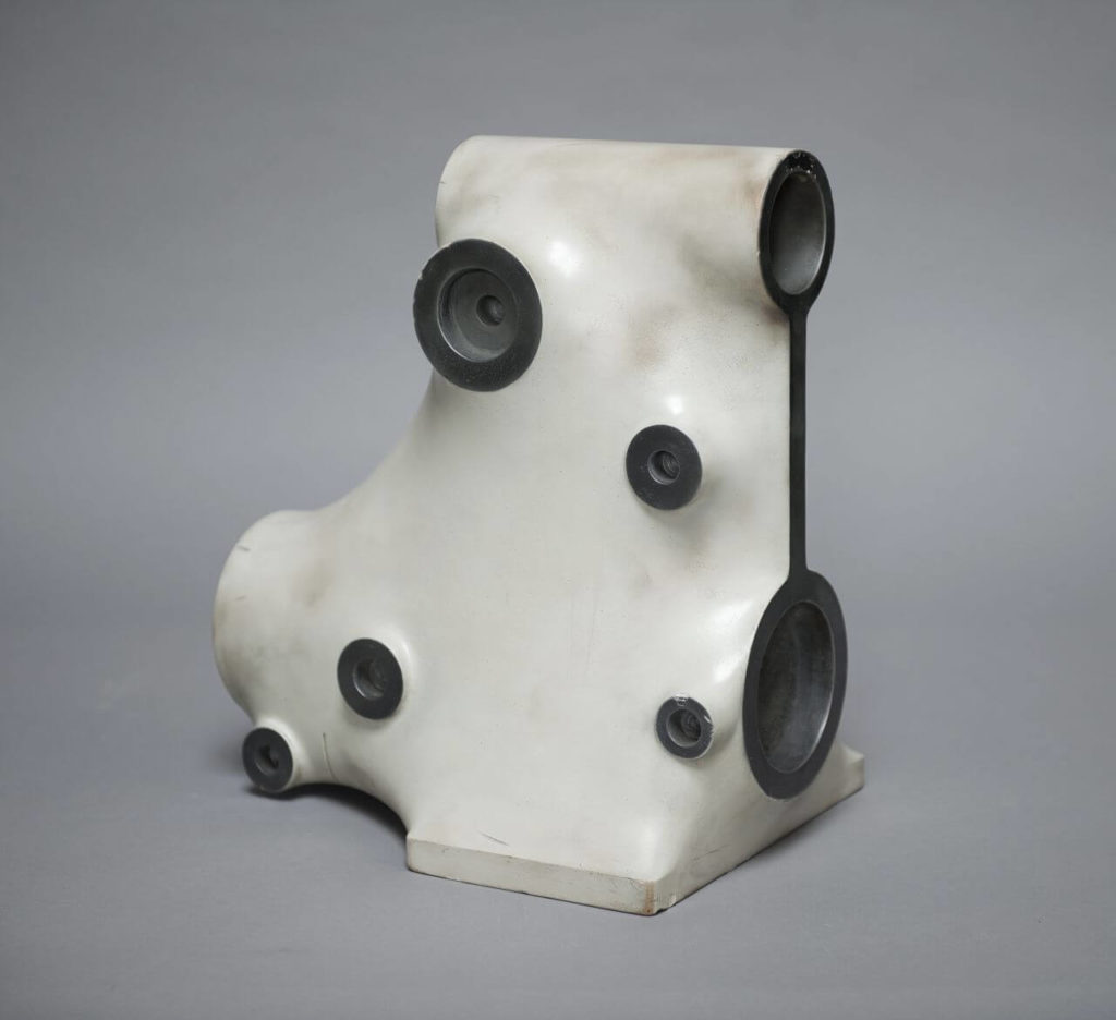 A white, almost abstract object with an organic shape, reminding a little of the body of an octopus. It has a solid base and is filled with many black round holes.