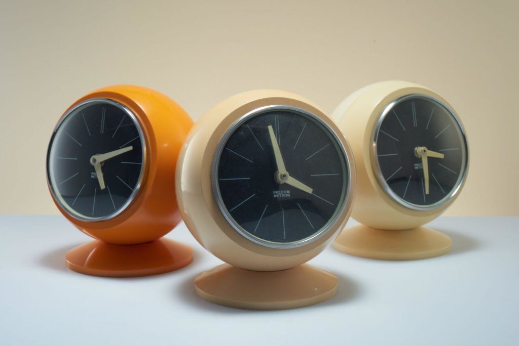 Three solid, thick wooden clocks. One is orange and two are beige. Their form is a ball-like sphere placed on a circular base. The hands of the clock are white against a black dial. No numerals, only white stripes in place of the hours.