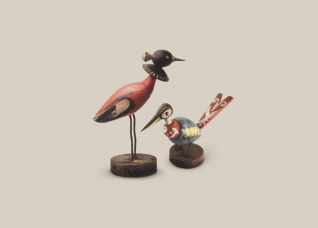 Two wooden figurines of little birds. Each is on a brown wooden base. The bird on the left has a little black head, a red body and black wings and thin long legs. The one on the right has a brown and white head, brown eyes, a black and golden beak, a red and blue body, and wings with yellow stripes. It has short thin legs. Its tail is red and decorated with a white folk-inspired pattern.
