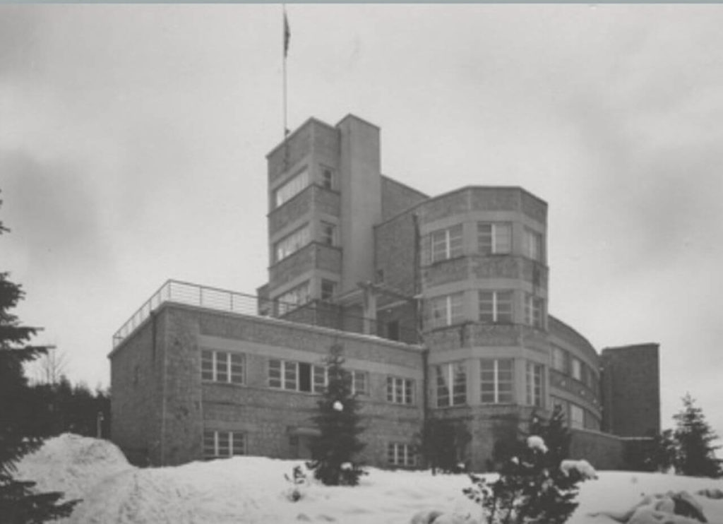 "A winter black and white shot with a lot of snow visible. It's a general view of a flat-roofed modernist mansion. The structure is enormous. The left half has two stories, the right three, and the middle area has up to five. There is a tall mast on top of the topmost roof with a folded flag on it. "