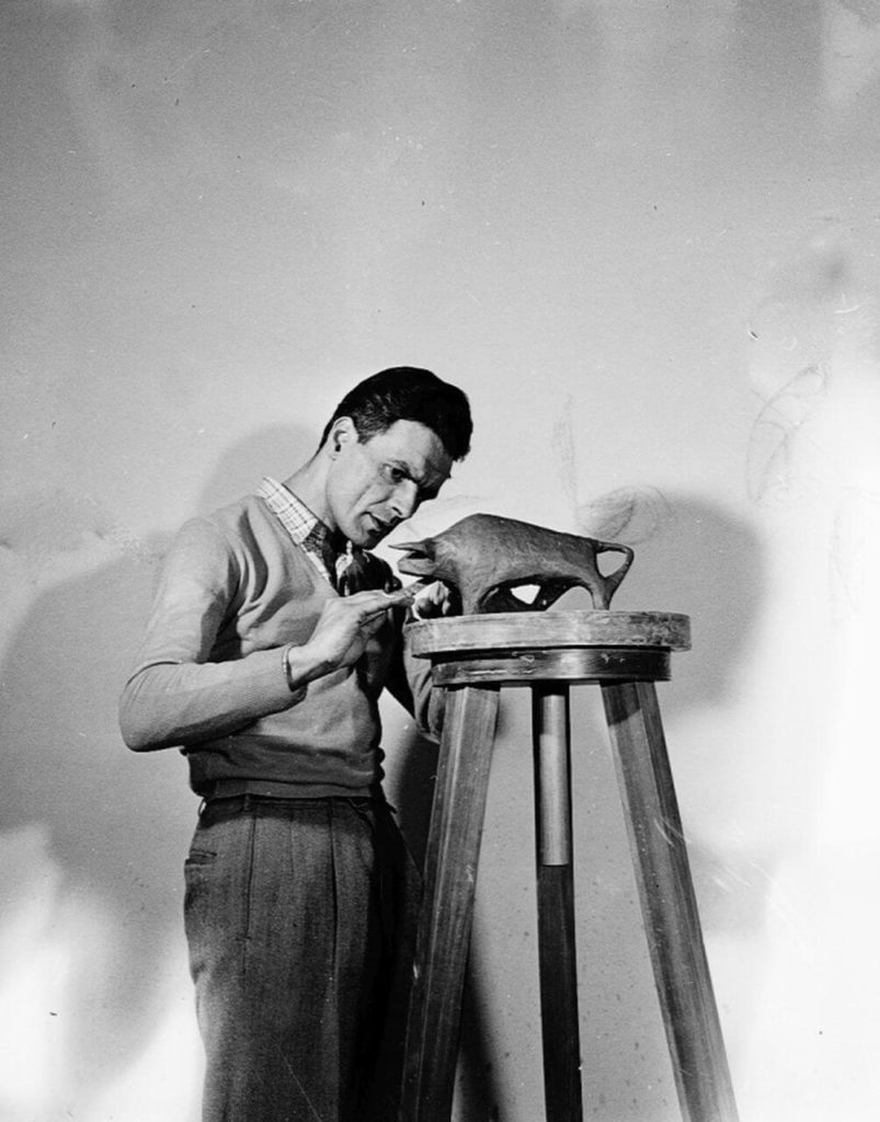 A black-and-white snapshot of a black-haired man meticulously constructing a little bull sculpture on a stool.