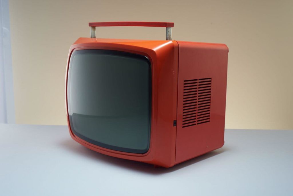 An old-style portable TV set. Its housing has a bright-red colour, and the screen seems grey. There are vents visible on the right side. At the top, there is a retractable handle. The screen part is a little rounded on the edges, while the back part is square.
