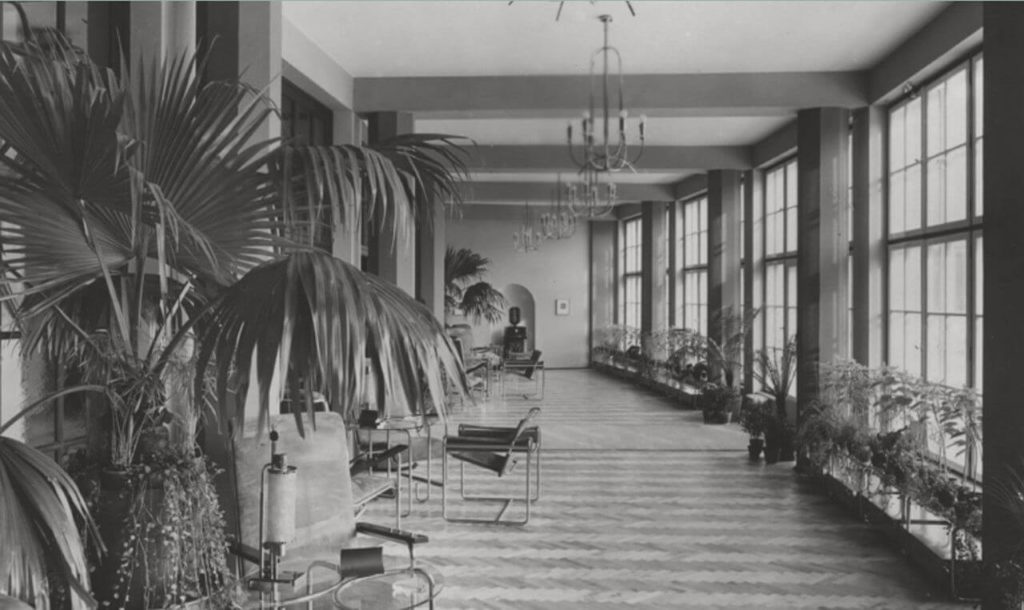 A black and white photograph depicting the interior of a modernistic building. The corridor is lengthy and there are many big flowers on the left side. Next to them are round transparent tables accompanied by minimalistic chairs. On the right side are large windows and rows of smaller flowers. Simple chandeliers are hanging from the ceiling.