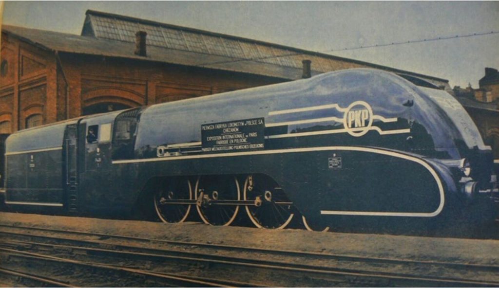 A colourized shot shows a large dark-blue steam locomotive from the side. It has golden lines and the PKP logo on it. The wheels are partially hidden by the body. The top of the front side is rounded.