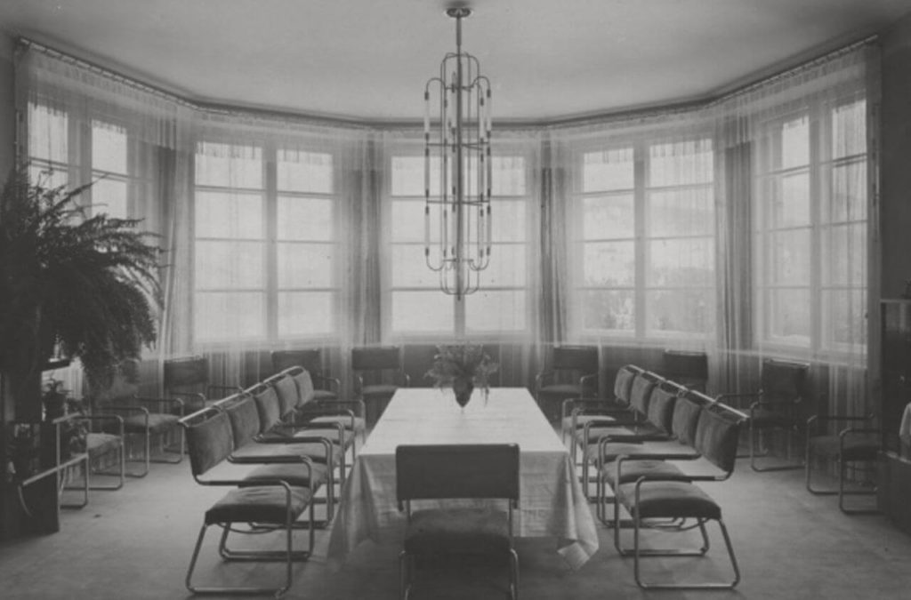 "A photograph shows the inside of Wisla Castle in black and white. The setting appears to be a conference room, complete with a large rectangular table draped in a white tablecloth and a vase in the center. On two sides of the table, there are rows of chairs and one chair at the end. "