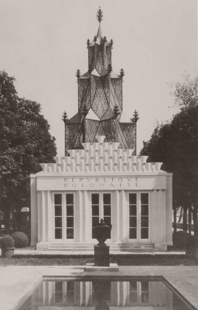 "An old monochromatic photograph. A white building with decorative, abstract motifs on the roof stands in the center. The structure has three large windows with the words 'REPUBLIQUE POLONAISE' above them. In front of the house, there is a pedestal with a vase on it, as well as a little square-shaped pond. A construction that resembles a soaring tower is located behind the building. "
