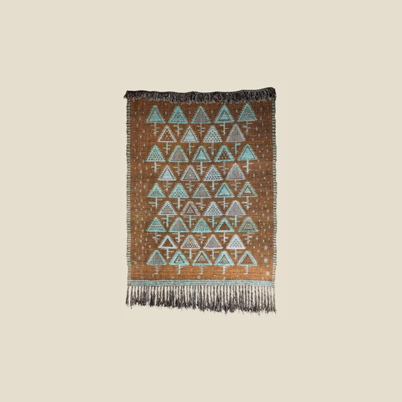 A brown fringy rug, decorated with a semi-regular ornament with geometrical, triangular, turquoise images of trees in the snow.