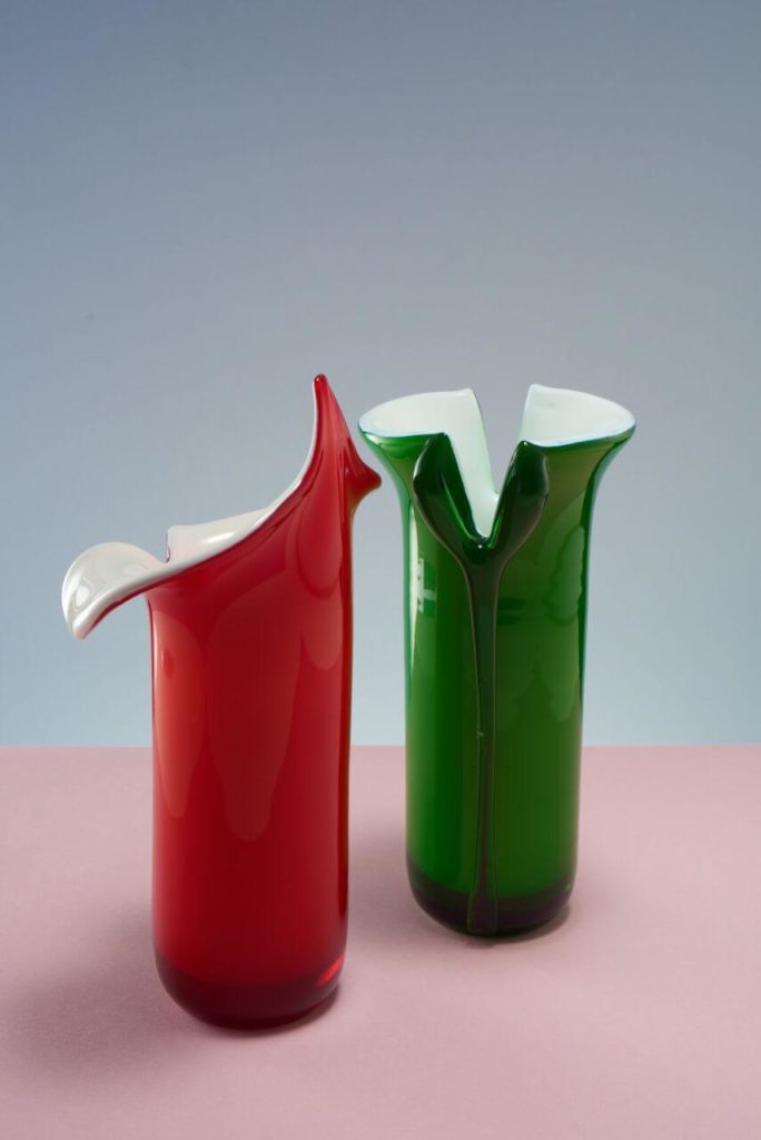 Two round vases. The vessel on the left is red, the right one is green, and both are white inside. The top edge of each looks like it is splitting, curling outwards.