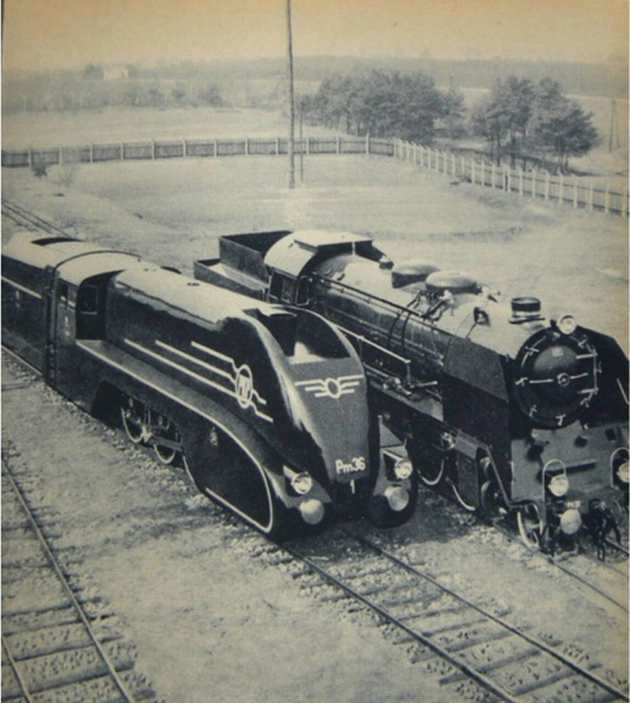 "Two steam locomotives are mounted on rails in this black and white image. The left one has a massive, rounded body, whereas the right one is smaller and has a cylinder-like front. "