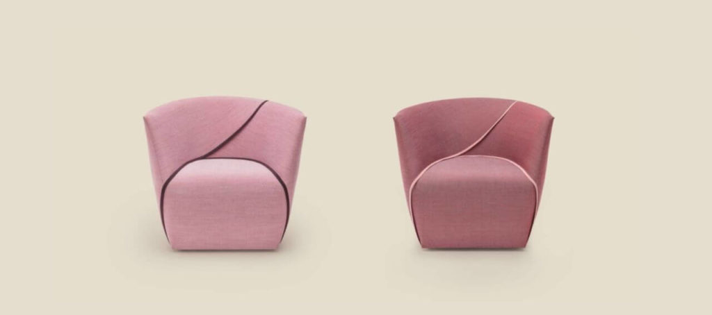Two comfortable armchairs of the same shape in different shades of salmon colour. Each armchair forms one solid structure without clearly separating the individual elements. Their silhouette is rounded, with slightly rounded backrests, through the centre of which runs a diagonal line connecting the sheets of fabric with each other. Another line surrounds the seat part.