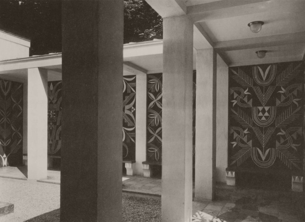 A monochromatic photograph showing the atrium of the Polish Pavilion. Its wall is filled with six sgraffito decorations, each depicting a simplified version of the coats of arms of major Polish cities placed among geometrical and stylized plant motifs.