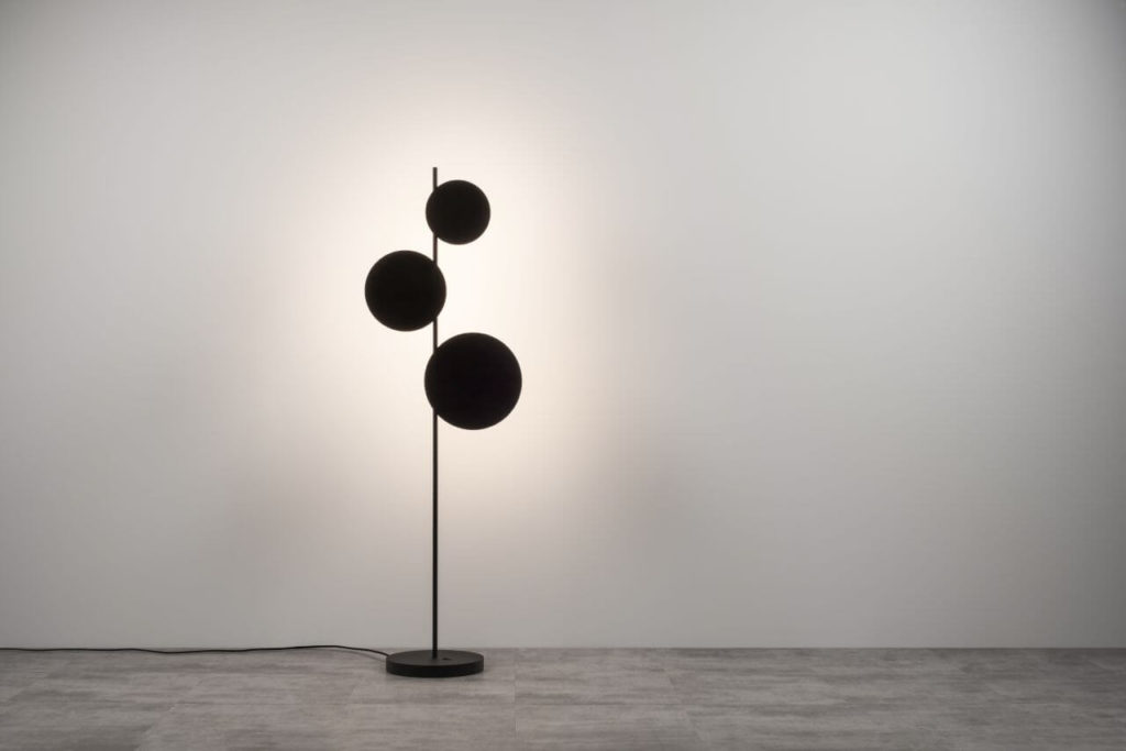A black floor lamp consisting of three round black discs on a vertical stand, facing the wall with their light source. The reflection on the wall gives the impression of a glow, and the discs resemble the sun during an eclipse.