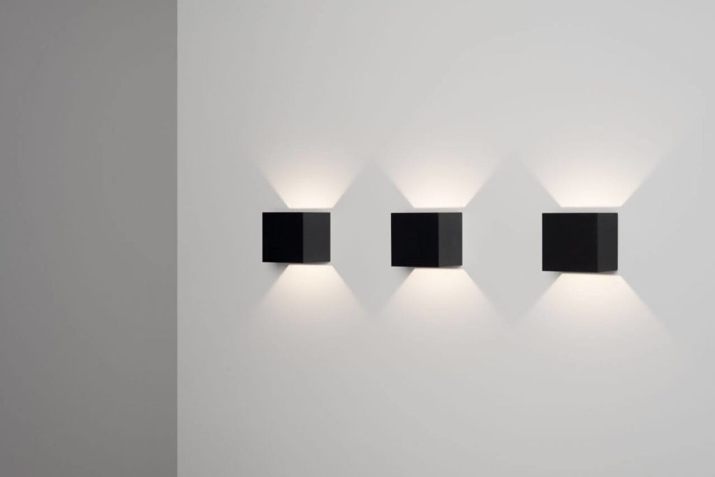 Wall lamp consisting of three black squares hung on a white wall. Each square creates a glow around itself caused by a hidden LED light source.