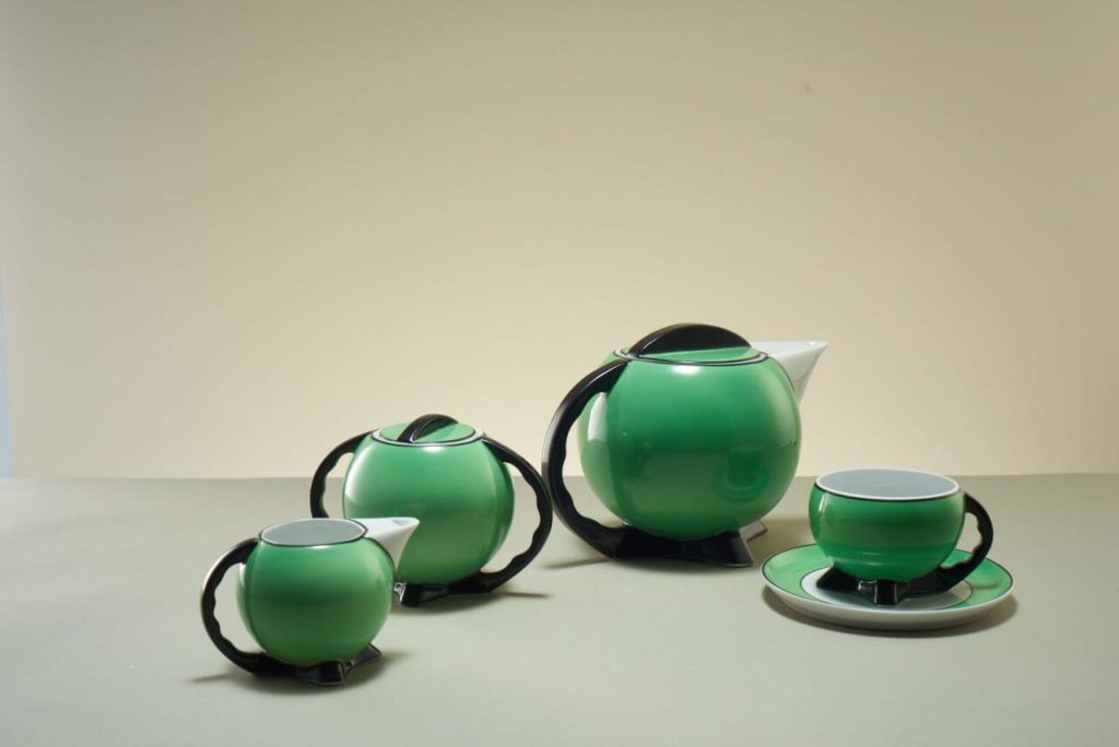 "A cofee set that includes a mug, sugar bowl, and cup with saucer. All of the objects are green on the outside, white on the interior, and have black handles that are stylized and slightly oversized. "