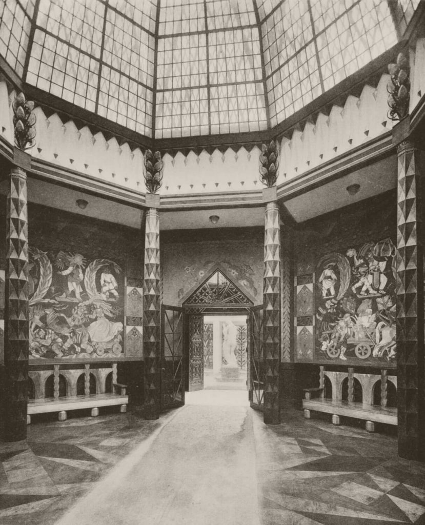 A black and white photograph of a fragment of the interior of the Polish Pavilion. The room has an octagonal shape. On each wall, there is an ornament by Zofia Stryjeńska. A piece of the dome is visible. Far in the background, there is a door leading outside, where the statue of a grieving woman stands.