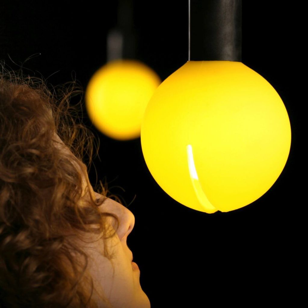 A photograph is a close-up of a red-haired woman's face looking very closely at a lamp that is hanging from a big black pipe. The lamp is round and looks like a glowing orange with a slit in the middle.