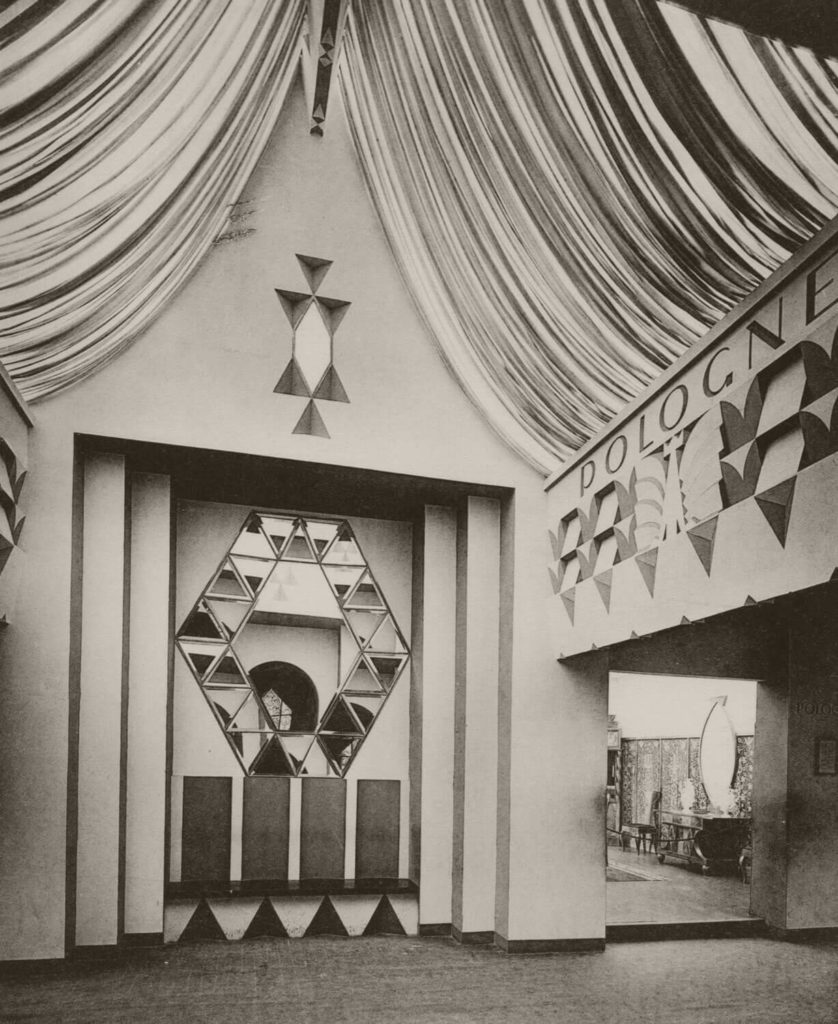 A black-and-white photograph of the Polish Pavilion's entryway and gallery. A wall with an artistic composition made of mirrors is in the picture's center. There is a passage on the right that leads to the other rooms. The simplified version of a white eagle with the text 'POLOGNE,' which means 'Poland' in French, may be found on the lintel above.