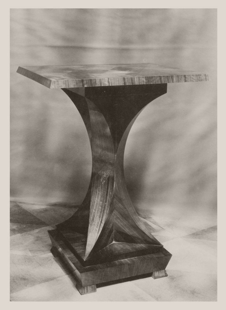 "A photograph of a solid-looking coffee table in black and white. It has a single thick X-shaped component that serves as a tabletop support. "