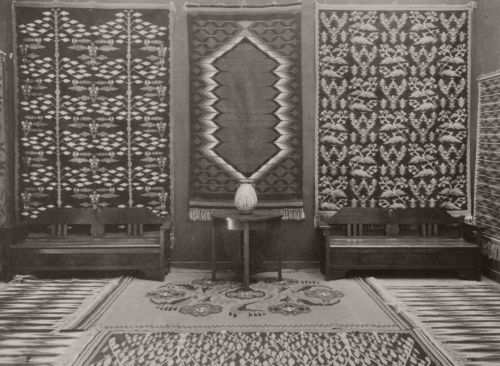 A black-and-white photograph of a room with Józef Czajkowski's ‘Kilim’ displayed in the centre. The work is placed between two other tapestries with flower motifs. Below, there is a small table with a vase on it.