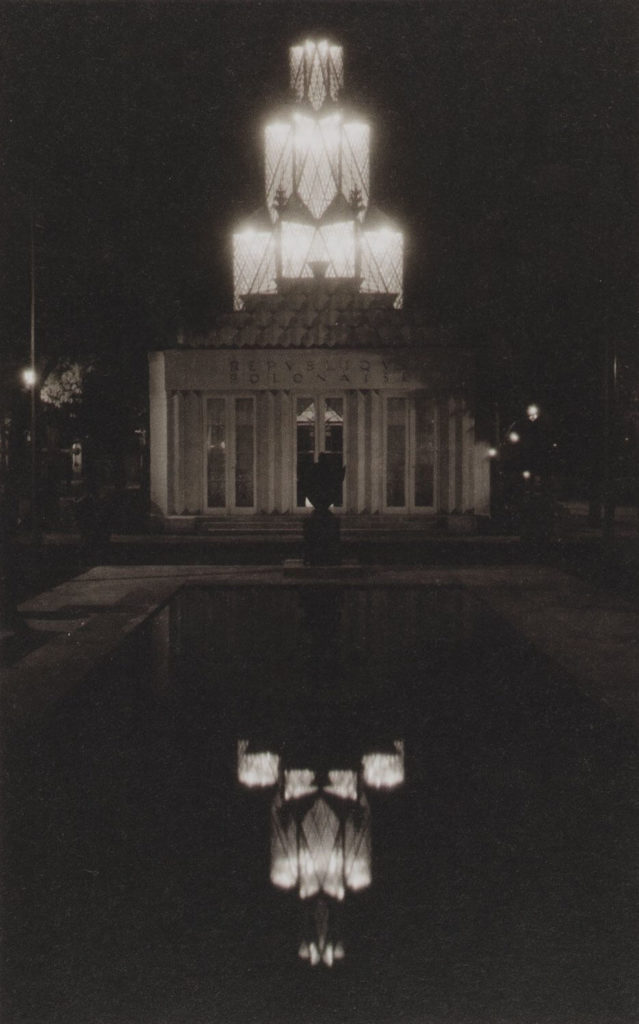 A monochrome shot of the Polish Pavilion taken at night from the outside. The white building's tower is now illuminated with lights. Their reflection can be seen in the pond's water.