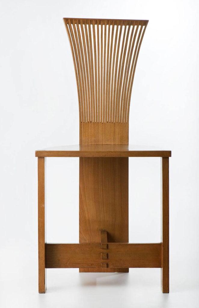 A wooden chair in a light brown color. It appears to be simple but stylized. The seat and front legs are standard, but the backrest, which also functions as a back leg, is tall, with a brush-like upper portion.