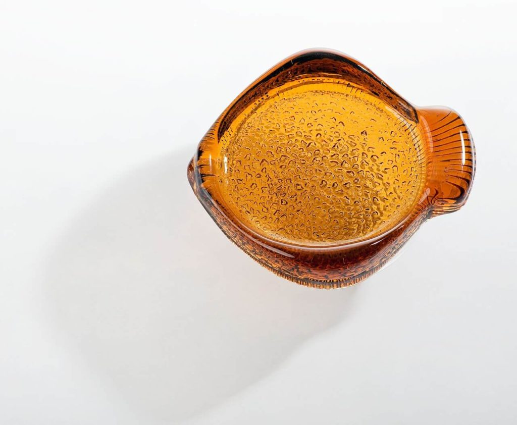 An aerial view of the glass semi-transparent amber-coloured ashtray, which is shaped like a fish. The main part of the vessel is a fish body, while the handle resembles a caudal fin. The surface of the object appears rough.