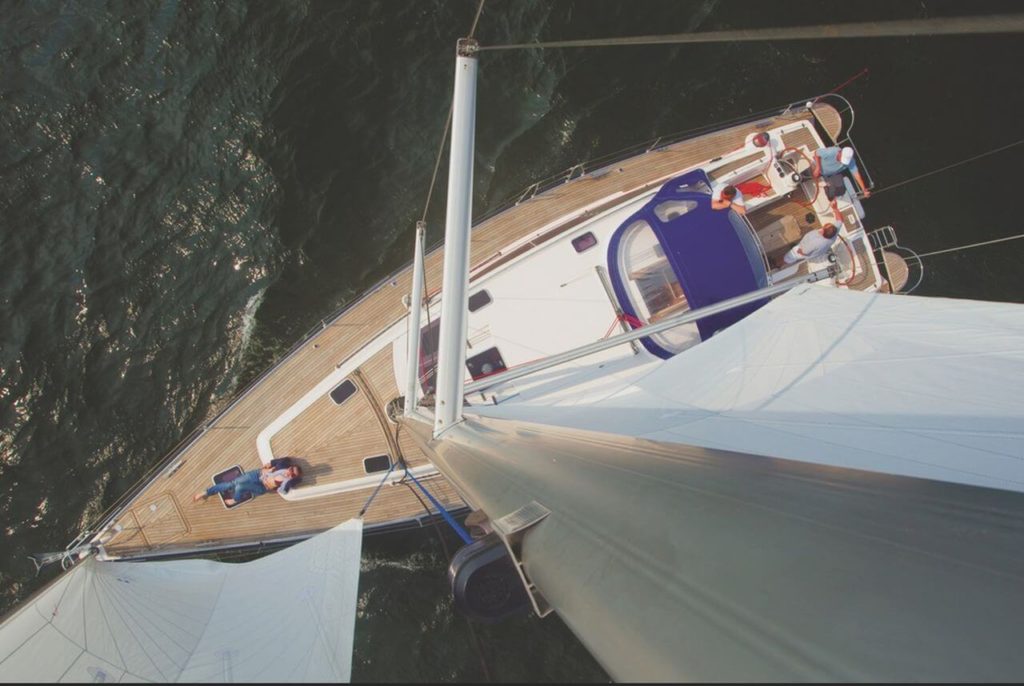 A sports yacht photographed from above, at mast height. Three men are standing at the steering wheels, talking. In front, by the bow, a fourth person is lying down and resting. The yacht is brown and white, and the roof of the deck cabin is purple.