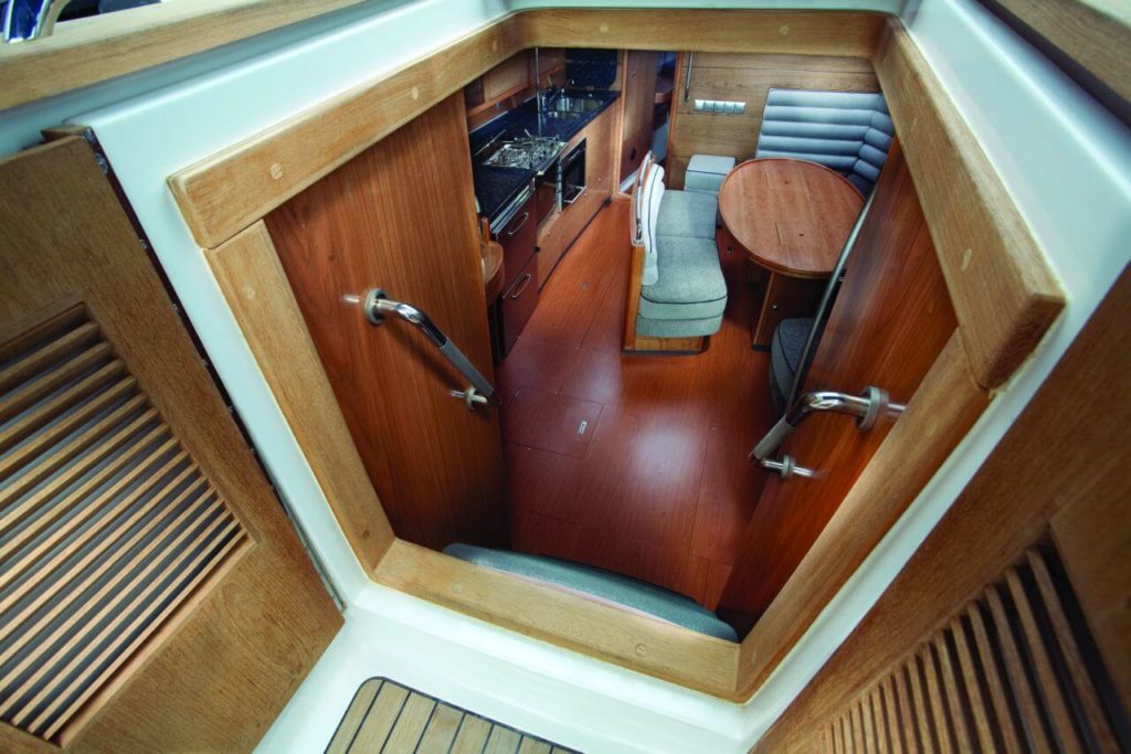 A luxurious cabin interior below the deck of a sports yacht. The walls and floor are wooden. You can see the kitchen, in the middle of which is placed a wooden table surrounded by grey-lined armchairs.