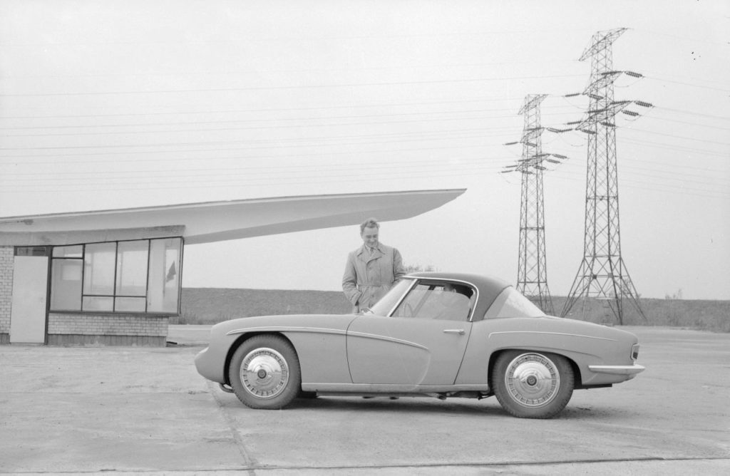 A black and white photograph. In the foreground, there is a two-door car with no backseats for the passengers. A middle-aged man is behind the wheel. Beside the car, another middle-aged man, wearing a trench coat, can be seen. Behind them is a modernistic little building with a prominent white roof. There are two transmission towers visible in the background.