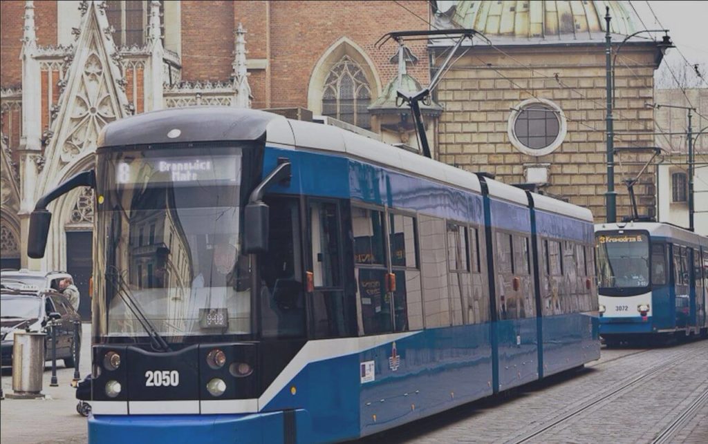 A colour photograph showing two modern trams. Both are blue with white accents, riding on the tracks in the centre of the old city. Above the windscreen, there is a display with the destination name. An historic church is visible in the background. The photo was taken in the city of Cracow.