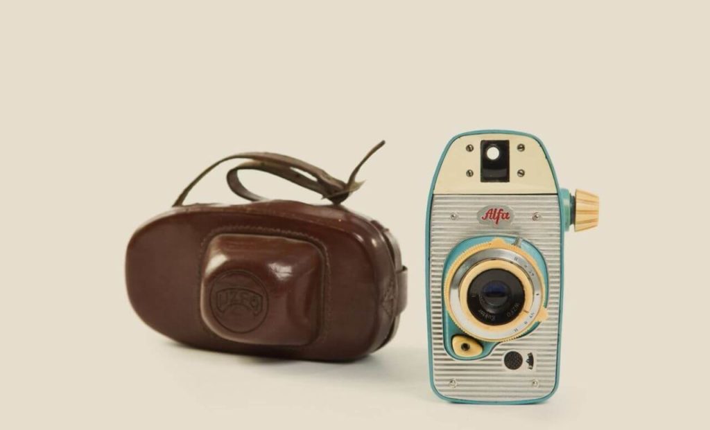 A small, brightly coloured analogue camera with curving corners that resembles a toy and a brown leather casing next to it. Grey, cream, and green dominate the camera, with yellow accents. It is vertically oriented. The lens is in the centre of the object, with a viewfinder above it and an ALFA red logo in between. The film advance lever is on the right side.