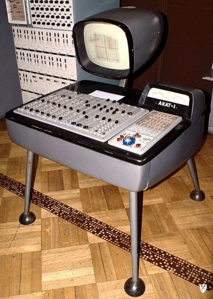 An old type of computer machine with many different elements combined into one unified body. It is grey and stands on four wide-spread legs. The lower panel has many levers, knobs, and gauges of various colours and shapes. The white writing says' Akat-I'. Above the panel, there is a relatively small screen, curvy on the edges.