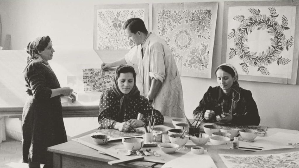 A black and white photograph depicts three women and a man in a room that looks like a painting studio. The women are located by the table full of painting accessories. The man behind them is putting a decorative image on the drawing board. Similar images are hanging on the walls.