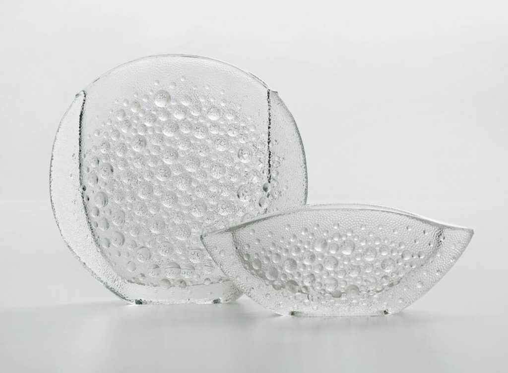 Two round glass vases, with the left one being a little taller. They have no colour and are semi-transparent. The outer surfaces of both are relief-finished. The convex pattern of glass bubbles blends in with the shape of the utensils in an optical illusion—larger bubbles in the protrusions, smaller ones along the farther edges.