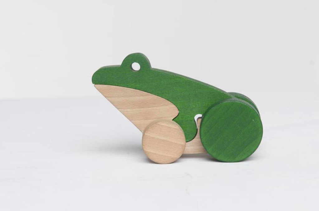A small wooden toy in the shape of a frog. It consists of two parts. The lower one is the underbelly of the frog, made of unpainted wood. The upper part, including the frog's head, is green. The toy can ride on two attached wheels.